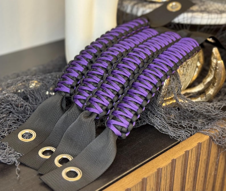 May include: Purple and black paracord wrist straps with metal rings. The straps are woven together in a complex pattern.