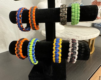 Puede incluir: Un expositor negro con ocho pulseras de paracord de colores. Las pulseras están hechas con diferentes combinaciones de colores, incluyendo negro, naranja, azul, verde, amarillo y gris.