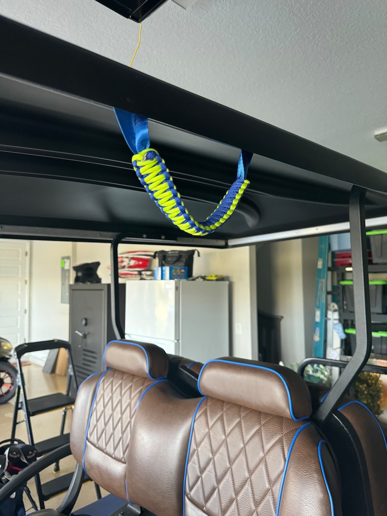 May include: A green and blue paracord handle hanging from the ceiling of a golf cart. The handle is made of two different colors of paracord, and it is tied in a knot at the top. The handle is hanging from the ceiling of a golf cart, and it is in focus.