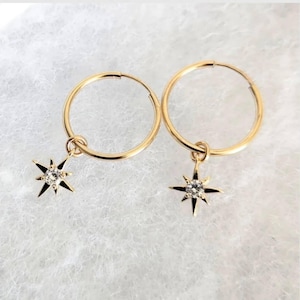 May include: A pair of gold-coloured hoop earrings. Each earring features a star-shaped charm with a central clear stone. The earrings are presented on a white, textured surface.