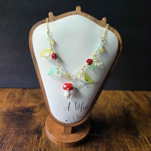 May include: A gold-tone chain necklace featuring a central red and white mushroom charm, accented with small red rose beads, translucent pale green leaf charms, and iridescent white spherical beads arranged symmetrically along the delicate metal links.