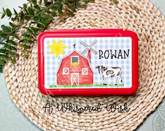 May include: Red lunchbox with a farm scene featuring a red barn, windmill, sun, and cow. The lunchbox has a blue and white checkered background and the name "ROWAN" is printed on the front. The lunchbox is made by "A Whispered Wish".