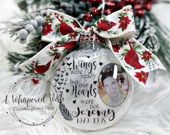 Puede incluir: Un adorno de Navidad de vidrio blanco con una foto de un hombre y el texto "Your wings were ready but our hearts were not Jeremy 1979-1998". El adorno está decorado con una cinta blanca con cardenales rojos y poinsettias.