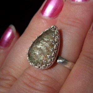 May include: A silver ring with a teardrop-shaped green and brown stone set in a silver bezel. The stone has a sparkly, iridescent finish.