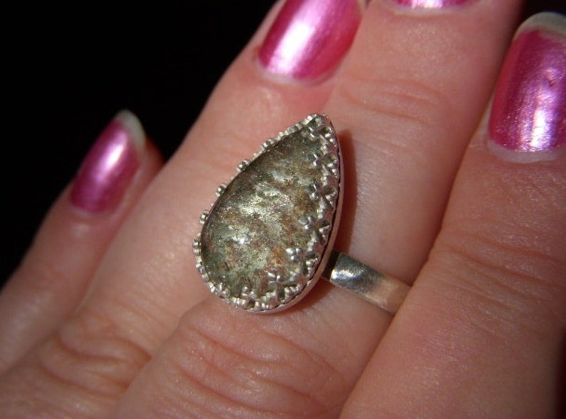 May include: A silver ring with a teardrop-shaped green and brown stone set in a silver bezel. The stone has a sparkly, iridescent finish.