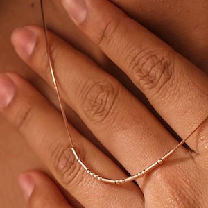 May include: A delicate rose gold necklace with a series of small, cylindrical and spherical beads. The necklace is held by a hand, showcasing the fine details of the jewelry. The beads are evenly spaced along the thin chain.