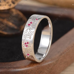 以下が含まれることがあります： ヒイラギとベリーのデザインが施されたシルバーリング。リングには、彫刻されたヒイラギの葉と、ベリーを表す小さな赤い宝石が特徴です。リングは木製の表面に展示されており、ジュエリーの複雑なディテールが際立っています。