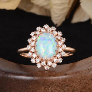 May include: A rose gold ring featuring a large oval opal gemstone surrounded by a halo of small, sparkling diamonds. The opal has a milky, iridescent appearance. The ring is set on a dark wooden surface.