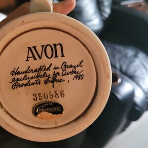 Puede incluir: Una taza de cerámica blanca con un asa marrón. La taza tiene la palabra "AVON" escrita en ella en letras negras. La taza también tiene el texto "Handcrafted in Brazil exclusively for Avon Products Inc., 1980" escrito en cursiva negra. La taza tiene una pegatina dorada con el texto "MADE IN GERMANY". La taza tiene el número "350586" escrito en ella en letras negras.