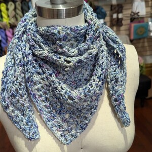 Könnte beinhalten: Ein gehäkelter Schal in Blautönen, Weiß und Rosa. Der Schal ist mit einer zarten Lochmuster-Masche gearbeitet und hat eine dreieckige Form.