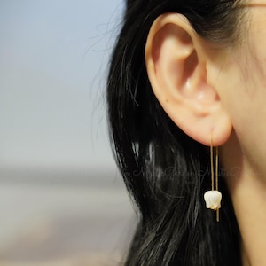 May include: A pair of delicate gold chain earrings with a white flower charm hanging from each chain. The earrings are shown on a person's ear.