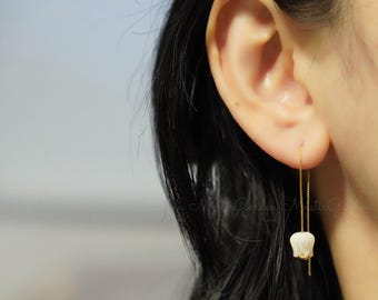 May include: A pair of delicate gold chain earrings with a white flower charm hanging from each chain. The earrings are shown on a person's ear.