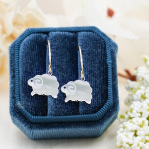 May include: A pair of gold hoop earrings with white sheep charms. The sheep are made of mother of pearl and have a detailed design.