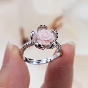 Peut inclure: Une bague en argent avec une bande délicate en forme de brindille et un motif de rose rose. La rose est sculptée dans la coquille et a un aspect réaliste détaillé.