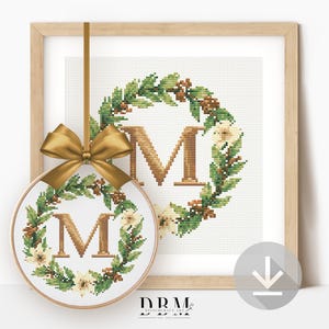 May include: A framed cross-stitch design featuring the letter "M" within a wreath of green leaves, white flowers, and brown accents. A smaller, circular cross-stitch with the same design hangs in front, adorned with a gold bow. The design is set against a white background.