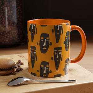 May include: A ceramic mug with a vibrant orange exterior and interior, featuring a repeating pattern of stylized dark gray faces with simple geometric features. The mug has a matching orange handle and a smooth, glossy finish throughout the surface.