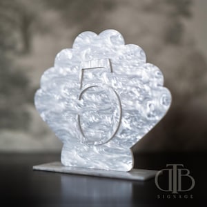 Puede incluir: Un número de mesa decorativo cinco, con forma de concha, hecho de acrílico veteado blanco y gris. El número está recortado, revelando el fondo. La base es un bloque rectangular. El logotipo "TB Signage" está en la esquina inferior derecha.