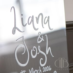 May include: A mirrored silver sign featuring the names "Liana & Josh" in cursive lettering. The date "21 March 2026" is also displayed. The sign has a reflective surface, suitable for weddings or special occasions.