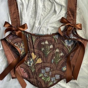 May include: A brown corset top with floral tapestry fabric and brown satin straps. The corset features a sweetheart neckline and is adorned with brown satin bows at the shoulders. The fabric has a pattern of flowers in shades of blue, yellow, and white.