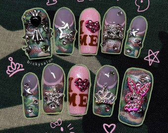 May include: Close-up of ten manicured fingernails with various designs. The nails are painted in shades of pink, green, and white. Some nails feature rhinestones, stars, and a crown. One nail has a pink Playboy bunny design. The nails are set against a camouflage background.