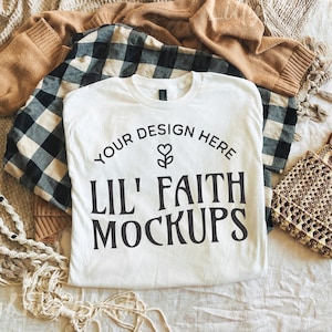 May include: A flat lay composition with a white t-shirt bearing the text "YOUR DESIGN HERE LIL' FAITH MOCKUPS", a tan sweater, a black and white checked shirt, a macrame necklace, and a small woven handbag with wooden handles.
