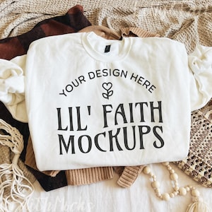 May include: A white crewneck sweatshirt with the text "YOUR DESIGN HERE" and "LIL' FAITH MOCKUPS" in black. The sweatshirt is surrounded by a brown and black plaid sweater, a tan sweater, a macrame bag, and a beaded necklace.