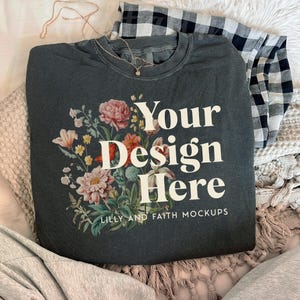 Puede incluir: Camiseta gris oscuro con el texto "Your Design Here" y un diseño floral. La camiseta se muestra en una cama con una camisa a cuadros en blanco y negro y un collar.