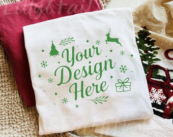 Könnte beinhalten: Weißes T-Shirt mit grünem Text „Your Design Here“ und festlichen Grafiken. Ein rotes T-Shirt ist hinter dem weißen Shirt zu sehen. Ein weihnachtliches Geschirrtuch befindet sich im Hintergrund.