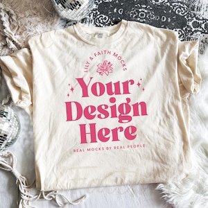 Puede incluir: Camiseta color crema con el texto rosa "Your Design Here", rodeada de un diseño floral y las palabras "Lily & Faith Mocks" y "Real Mocks by Real People". La camiseta se muestra sobre una superficie blanca.