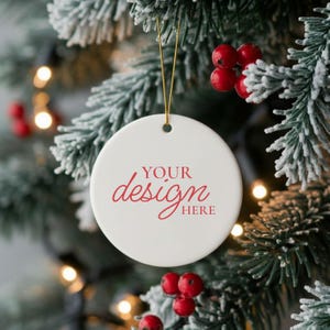 May include: A white, round Christmas ornament with the text "YOUR design HERE" in red script. The ornament hangs from a gold string against a backdrop of a snow-covered evergreen branch with red berries and twinkling lights.