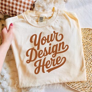 Könnte beinhalten: Cremefarbenes T-Shirt mit dem Text "Your Design Here" in brauner Schrift im Retro-Stil. Das Shirt wird auf einer weichen, strukturierten Oberfläche präsentiert, was auf eine bequeme Passform hindeutet. Das Etikett lautet "Comfort Colors".