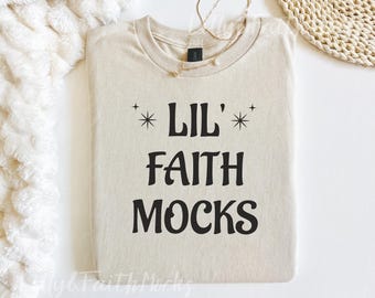 Puede incluir: Una camiseta beige con el texto "LIL' FAITH MOCKS" en negro, con diseños de estrellas arriba y abajo de la palabra "LIL'". Un collar dorado descansa sobre la camisa. La camisa está doblada cuidadosamente sobre una superficie blanca.