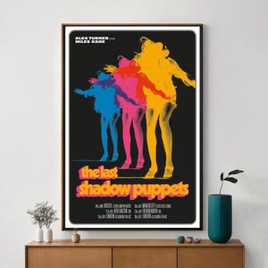 Peut inclure: A framed poster for "The Last Shadow Puppets" on a black background. It features three silhouettes in blue, pink, and yellow. The band's name is in pink and yellow, with tour dates below.