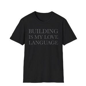 May include: A black short-sleeved t-shirt featuring the text "BUILDING IS MY LOVE LANGUAGE" printed in a dark grey, serif font across the center of the chest. The garment has a classic crew neckline and a standard fit with short sleeves.