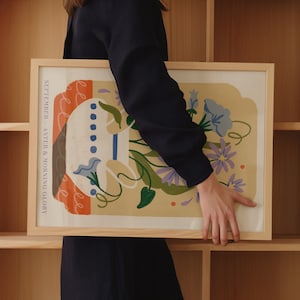 Op de afbeelding: Een ingelijste print met een lichtgele achtergrond met een gestileerde illustratie van een vaas met blauwe en paarse bloemen. De tekst "SEPTEMBER - ASTER & MORNING GLORY" staat gedrukt aan de linkerkant van de print.