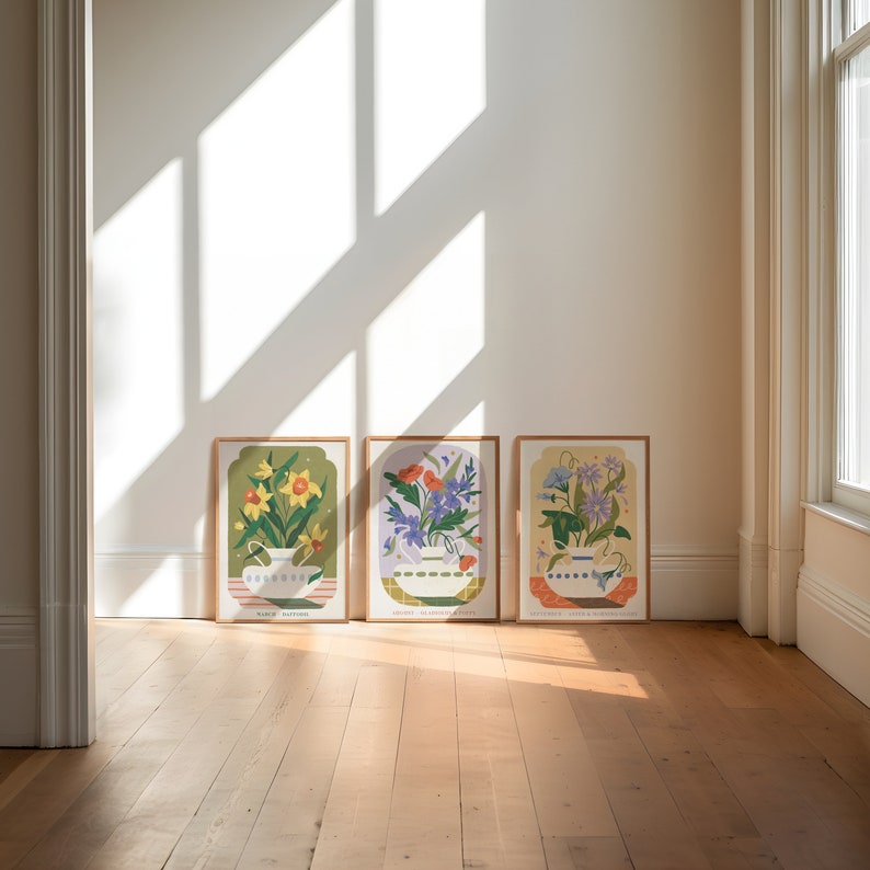 Op de afbeelding: Drie ingelijste prints met bloemillustraties. Elke print toont een ander type bloem in een witte kan. De prints zijn gelabeld met de maand en de naam van de bloem. De prints bevinden zich op een houten vloer.