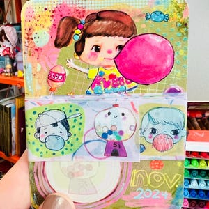 May include: A colorful notepad featuring a cartoon girl blowing a pink bubble. The notepad has a whimsical design with candy and bubblegum themes. The bottom section displays the text "nov 2024".