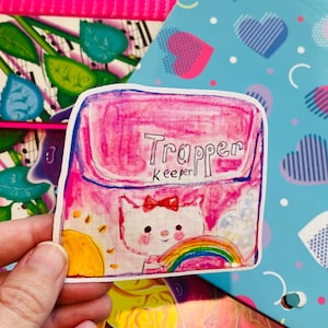 May include: A pink Trapper Keeper with a white cat and rainbow design. The text "Trapper Keeper" is written on the front of the binder.