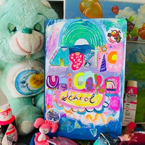 May include: A colorful painting on canvas with the words "Mystic Unicorn School" in a rainbow of colors. The painting is surrounded by toys and art supplies, including a blue teddy bear, a pink toy, and a paint roller.