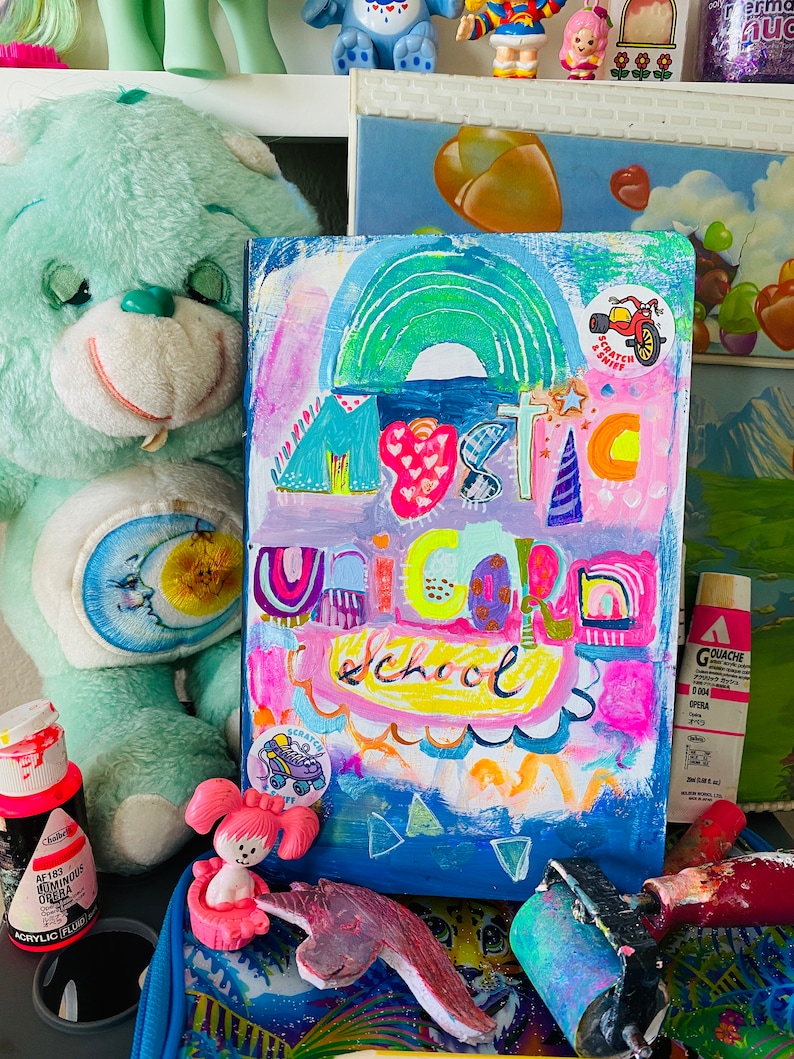 May include: A colorful painting on canvas with the words "Mystic Unicorn School" in a rainbow of colors. The painting is surrounded by toys and art supplies, including a blue teddy bear, a pink toy, and a paint roller.