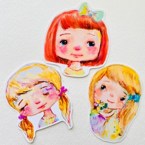 Puede incluir: Tres pegatinas troqueladas con ilustraciones de acuarela de niñas. Una tiene el pelo rojo y un lazo, otra tiene coletas rubias y la tercera tiene el pelo rubio y sostiene flores. Las pegatinas tienen un estilo artístico y caprichoso.