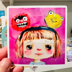 May include: Square sticker featuring a whimsical illustration of a girl with short red hair, a blue striped shirt, and rosy cheeks. A red monster and a yellow frog are on her head, set against a pink background.