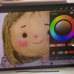 May include: A digital illustration of a girl's face on a tablet screen. The tablet is being used with a stylus to select colors from a color wheel on the screen. The color wheel has a variety of colors, including red, orange, yellow, green, blue, indigo, and violet. The girl's face is drawn in a cartoon style with large eyes and a wide smile.