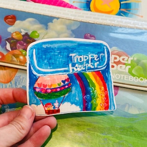 May include: A colorful sticker featuring a hot air balloon with a rainbow and the words "Trapper Keeper." The balloon has a basket with two cartoon characters. The sticker is blue, pink, red, yellow, and green.