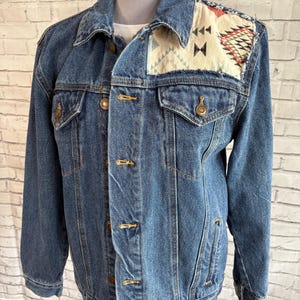 May include: A blue denim jacket with a decorative patch on the shoulder. The patch features geometric patterns in red, black, and cream colors. The jacket has a classic button-up design with two chest pockets.
