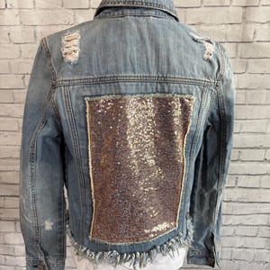 May include: Light blue denim jacket with distressed details and a large sequined panel on the back. The jacket has a frayed hem and a classic collar. The sequin panel is framed with a gold trim.