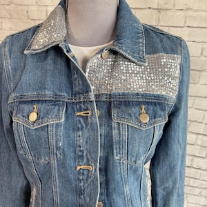 May include: A denim jacket with silver sequin embellishments on the collar and across the chest. The jacket has two front pockets and gold-colored buttons. The garment is displayed on a mannequin against a brick wall background.
