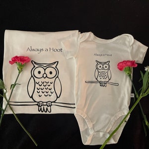 May include: Two white t-shirts with an owl graphic and the text "Always a Hoot". The shirts are on a black background with pink and white flowers.