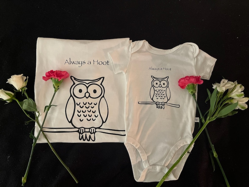 May include: Two white t-shirts with an owl graphic and the text "Always a Hoot". The shirts are on a black background with pink and white flowers.
