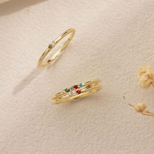 May include: Two gold rings featuring small, colorful gemstones. One ring has a single green stone and a clear stone, while the other ring features a row of five small stones in pink, green, blue, red, and clear colors set into the thin gold band.